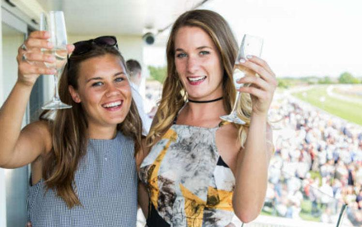 Two ladies holding champagne glasses while standing on a balcony at Bath Racecourse.